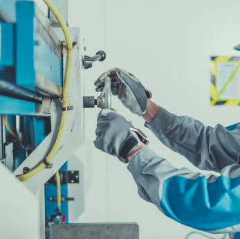 A technician in grey work gloves and a grey uniform uses a tool to work on industrial machinery in a