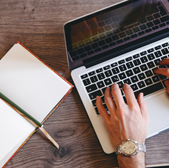 A person wearing a watch types on a laptop at a wooden desk, with an open notebook and pen beside th