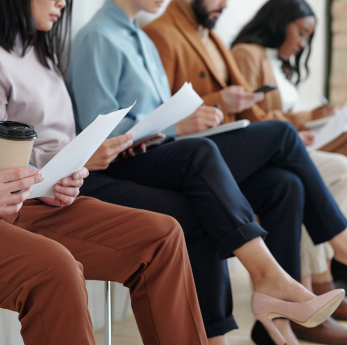 A row of smartly dressed people sit in a waiting area holding CVs and papers, suggesting a job inter