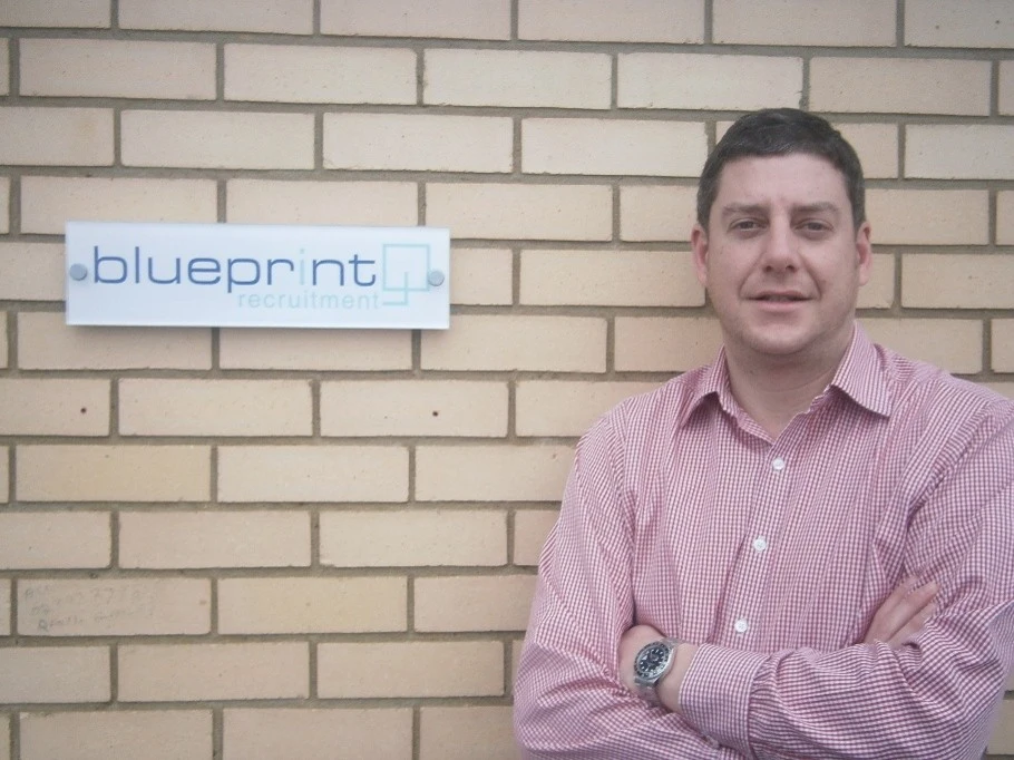 Mark Butter standing with his arms crossed beside an early Blueprint Recruitment sign on a brick wal